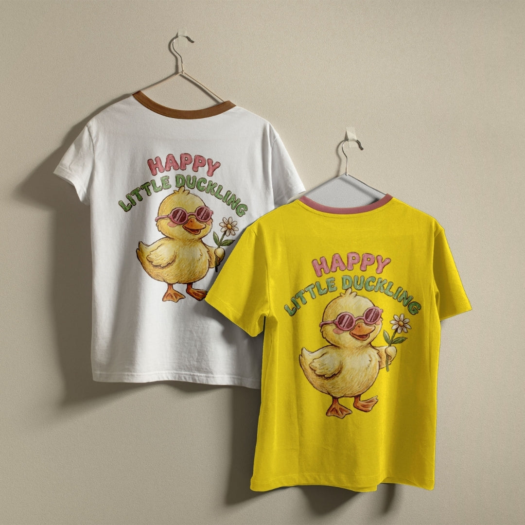 Two children's short-sleeve graphic T-shirts hanging on hangers against a plain beige wall. One shirt is white with a brown collar trim and a yellow duckling graphic with the text 'HAPPY LITTLE DUCKLING' in pink and green.