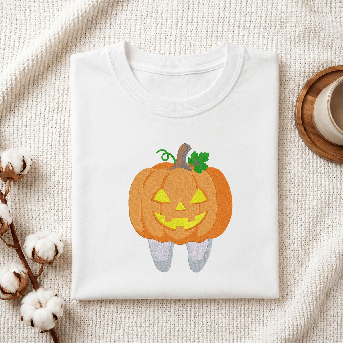 Folded white cotton t-shirt mockup featuring a cute jack-o-lantern pumpkin tooth Halloween dental machine embroidery design, styled with cotton stems and a wooden cup