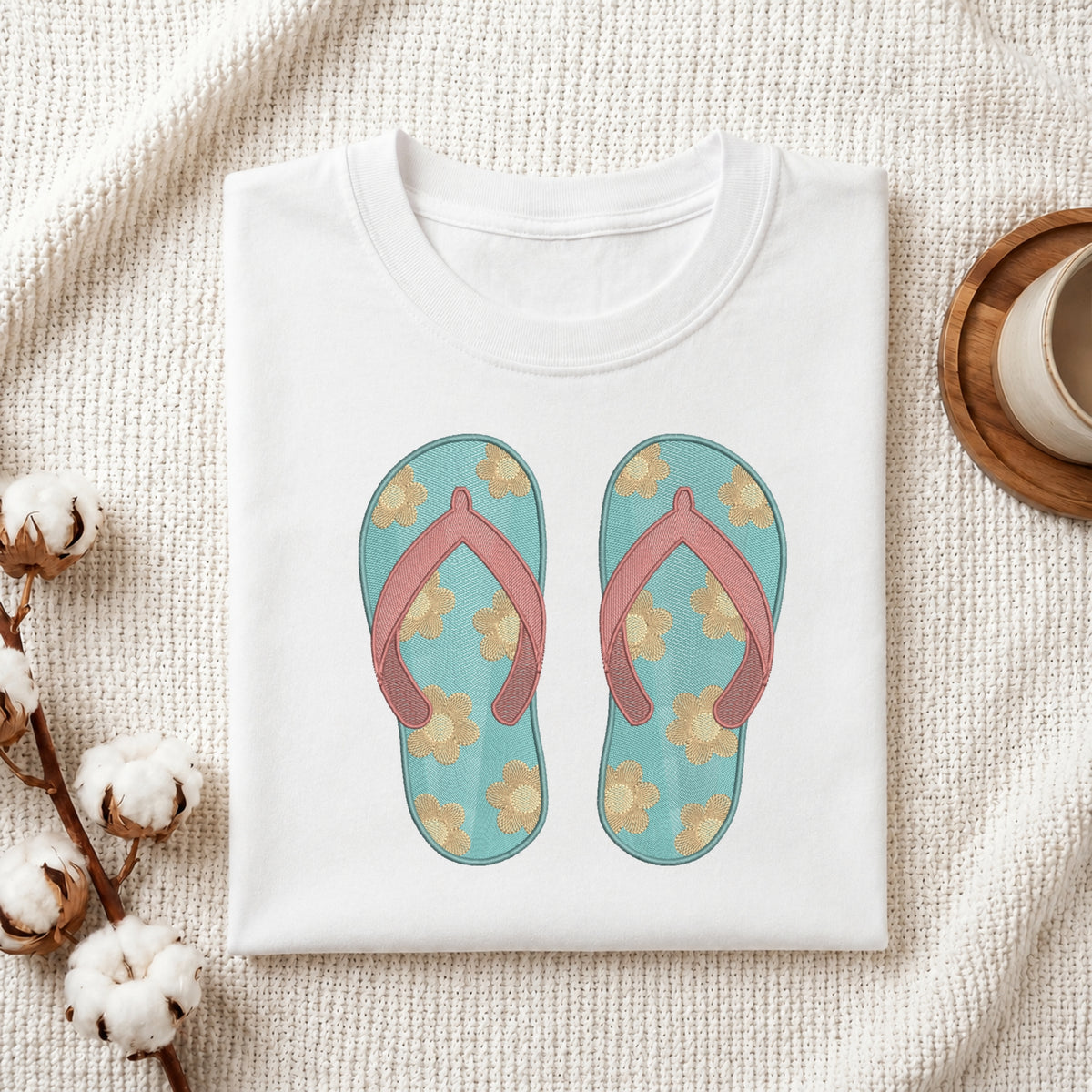 A folded white T-shirt laid flat on a textured cream fabric background, featuring a printed graphic of a pair of turquoise flip-flops with pink straps and beige floral motifs. Decorative cotton bolls and a small cup appear at the edges as styled props.