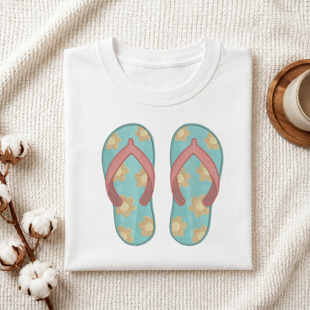 A folded white T-shirt laid flat on a textured cream fabric background, featuring a printed graphic of a pair of turquoise flip-flops with pink straps and beige floral motifs. Decorative cotton bolls and a small cup appear at the edges as styled props.