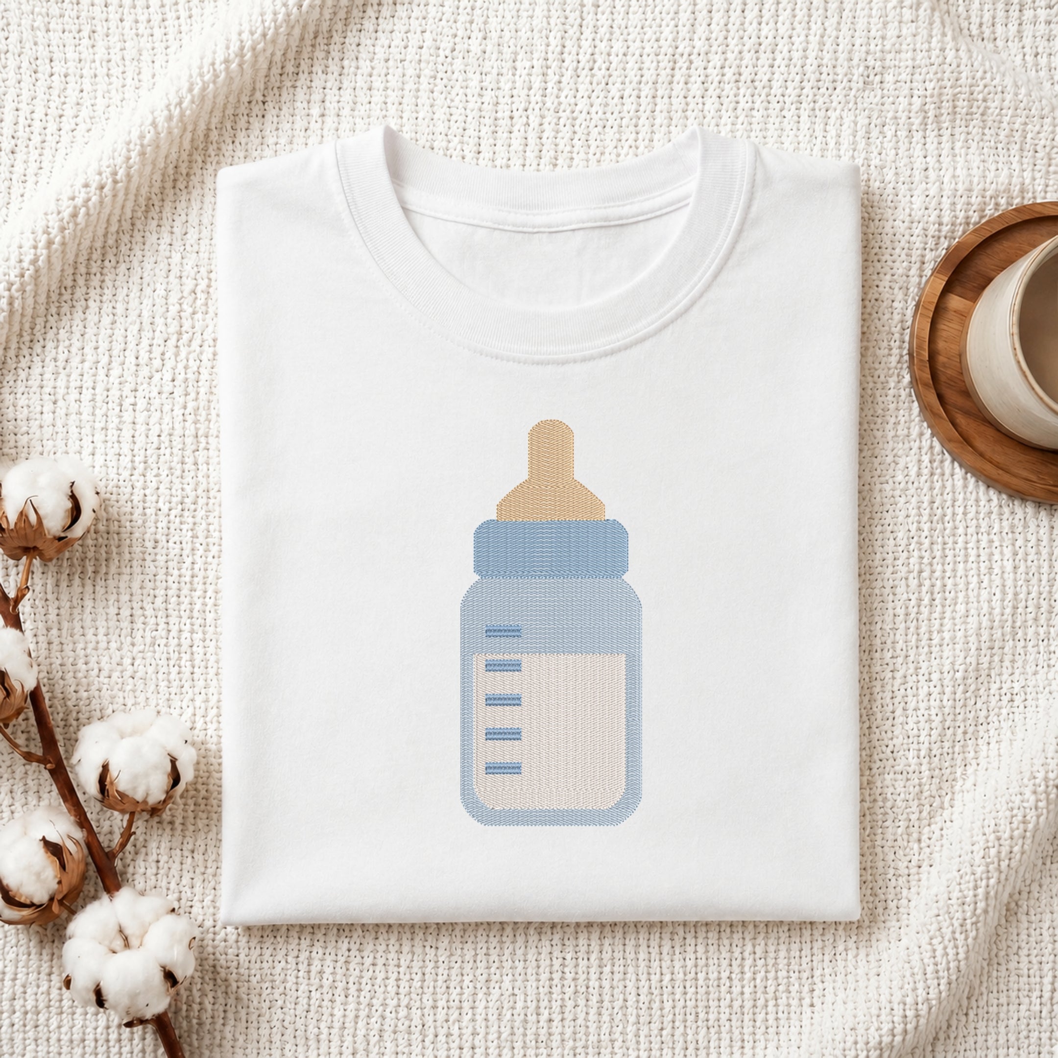 A folded white t-shirt with a baby bottle graphic, styled on a textured cream fabric with cotton bolls and a wooden cup.