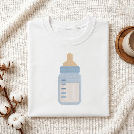 A folded white t-shirt with a baby bottle graphic, styled on a textured cream fabric with cotton bolls and a wooden cup.