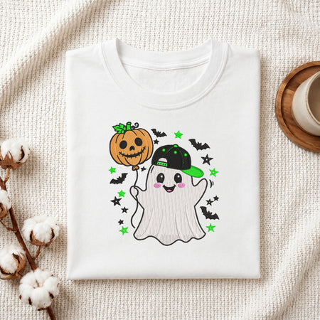 A neatly folded white t-shirt featuring the embroidered ghost in a baseball cap holding a pumpkin balloon graphic, displayed on a textured white knit background with cotton stems and a wooden cup.