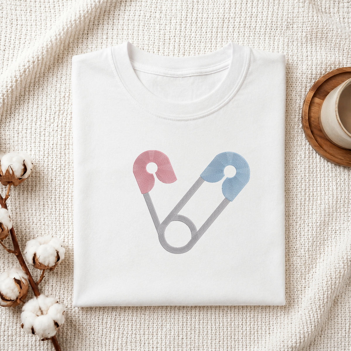 A folded white t-shirt with a baby safety pin graphic in pink, blue, and gray, styled on a textured cream knit blanket with cotton bolls and a cup in the background.
