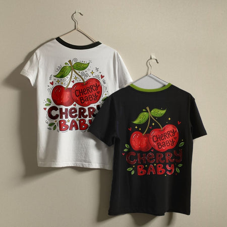Two graphic t-shirts hanging on hangers against a plain beige wall. The left shirt is white with a black collar and features a large back graphic of two red cherries with green leaves and the text 'CHERRY BABY'.