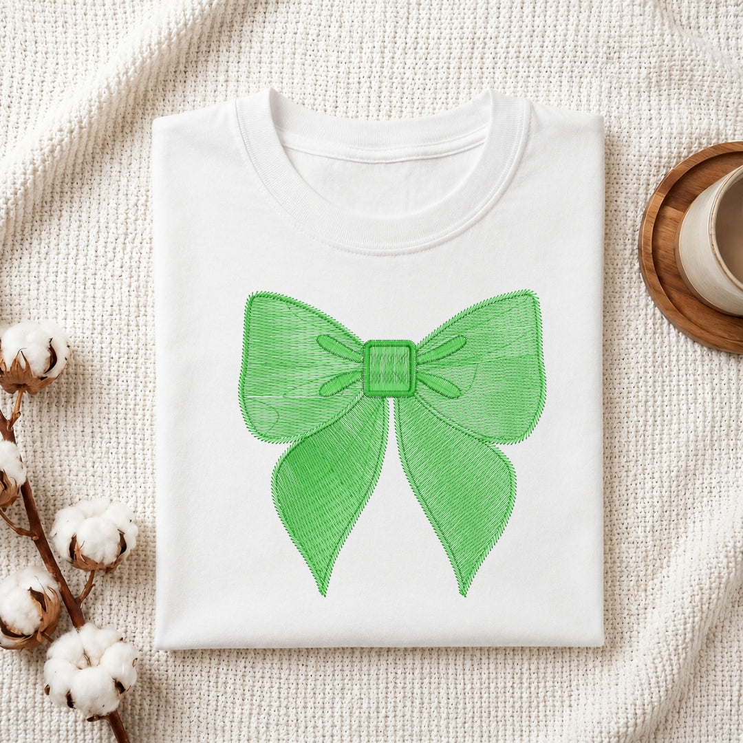 A folded white crewneck sweatshirt or t-shirt with a bright green bow graphic printed on the front, displayed on a textured cream knit blanket with cotton bolls and a cup in the background.