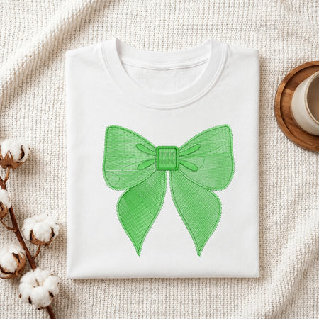 A folded white crewneck sweatshirt or t-shirt with a bright green bow graphic printed on the front, displayed on a textured cream knit blanket with cotton bolls and a cup in the background.