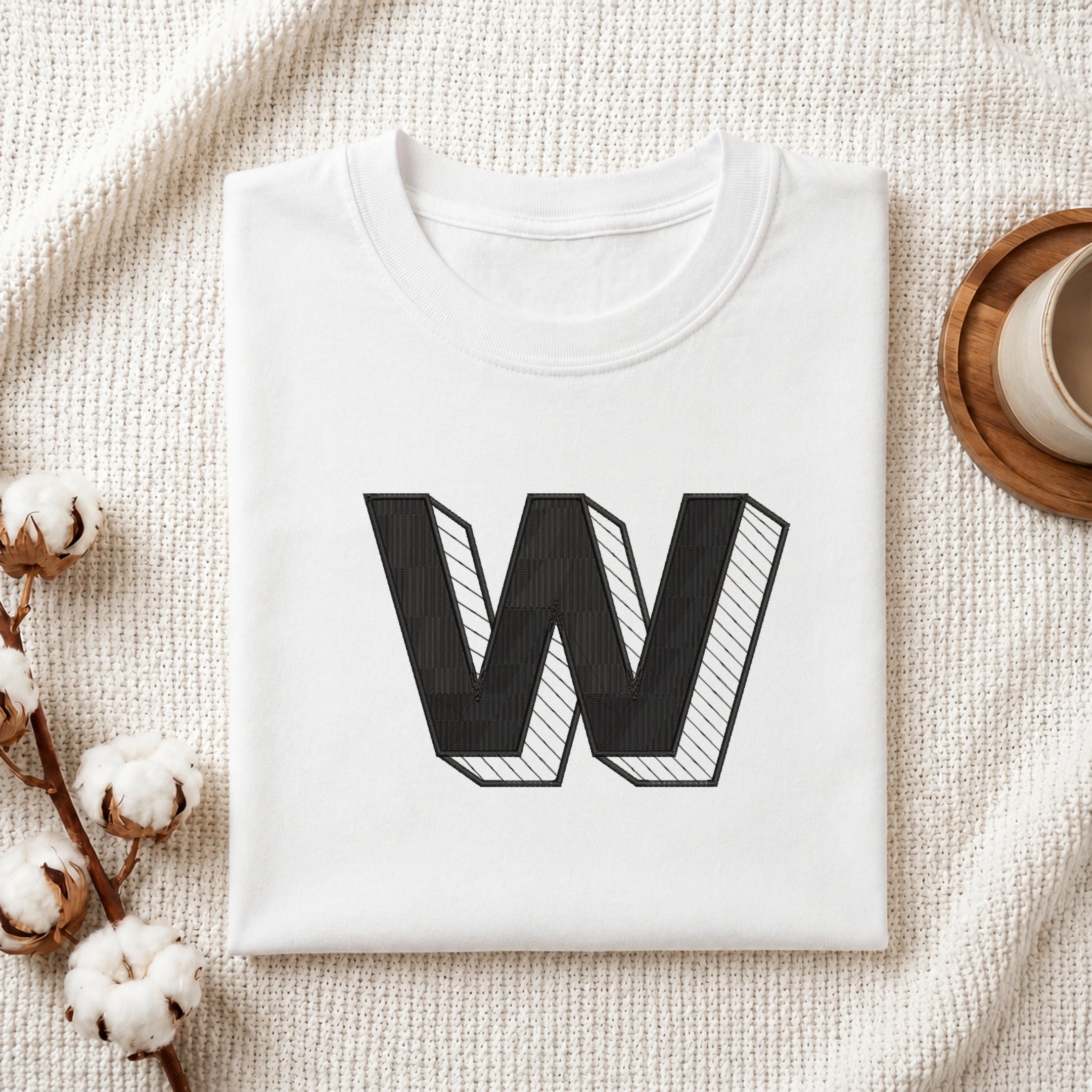 A folded white t-shirt with a large black block letter 'W' printed on the front, styled on a textured cream knit blanket with cotton bolls and a cup on a wooden tray.
