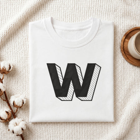 A folded white t-shirt with a large black block letter 'W' printed on the front, styled on a textured cream knit blanket with cotton bolls and a cup on a wooden tray.