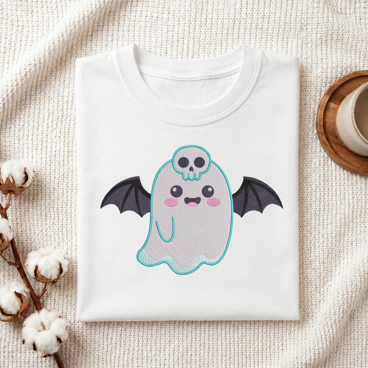A folded white t-shirt with a cute cartoon ghost graphic, styled on a textured cream fabric with cotton bolls and a coffee cup in the background.