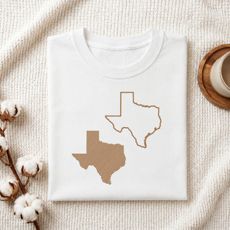 A folded white T-shirt laid flat on a textured cream fabric background, featuring a graphic of Texas in the shape of a kraft paper bag on the left and a white outline of Texas on the right. Styled flat-lay props include cotton bolls on the left and a cup on a wooden coaster on the right.