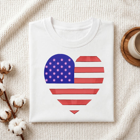 A folded white crewneck t-shirt featuring a heart-shaped American flag graphic on the front, styled on a textured cream fabric background with cotton bolls and a wooden cup.