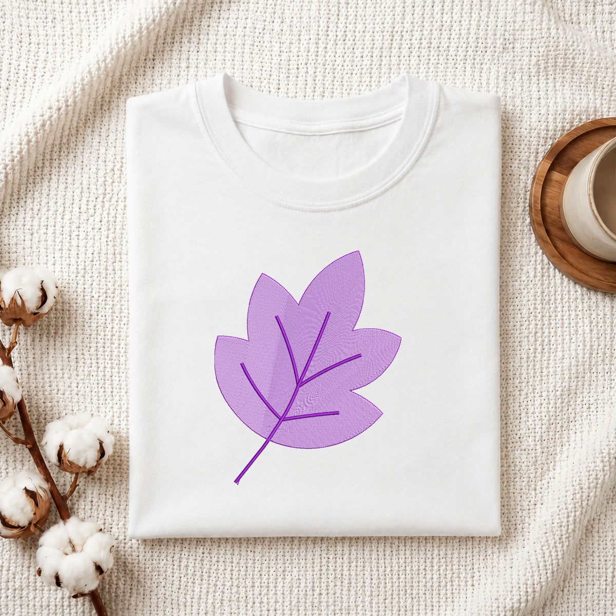 Folded white cotton t-shirt mockup featuring a purple autumn leaf machine embroidery design, styled with cotton stems and a wooden cup.