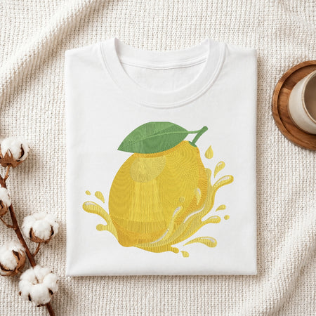 A white folded t-shirt featuring a detailed, yellow embroidered lemon with green leaves and a juice splash design.
