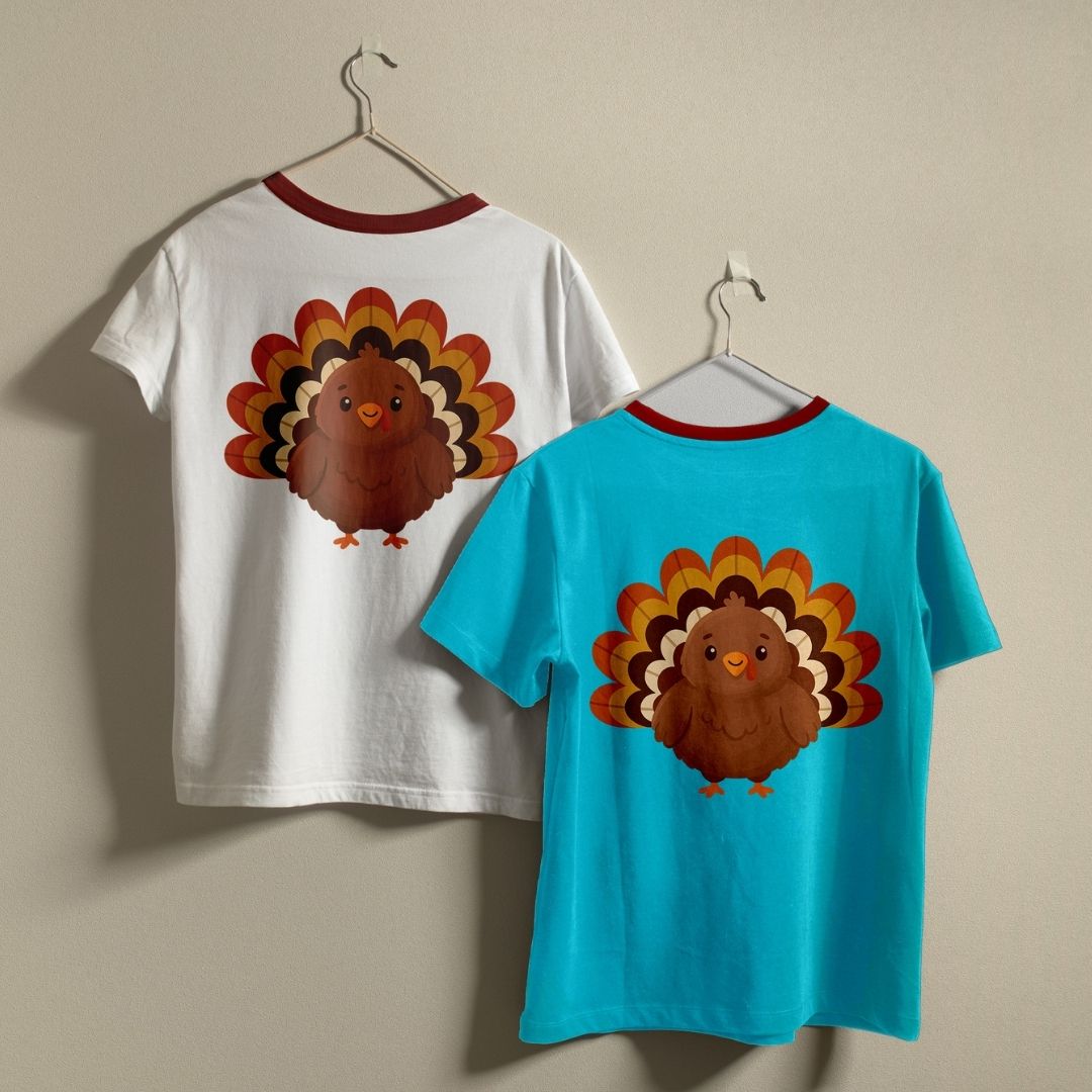 Two children's short-sleeve ringer T-shirts hanging on hangers against a plain beige wall. One shirt is white with a red collar trim and a large brown turkey graphic on the back.