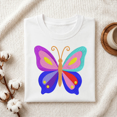 A folded white crewneck t-shirt featuring a colorful embroidered butterfly design, styled on a textured cream fabric background with cotton bolls and a wooden cup.
