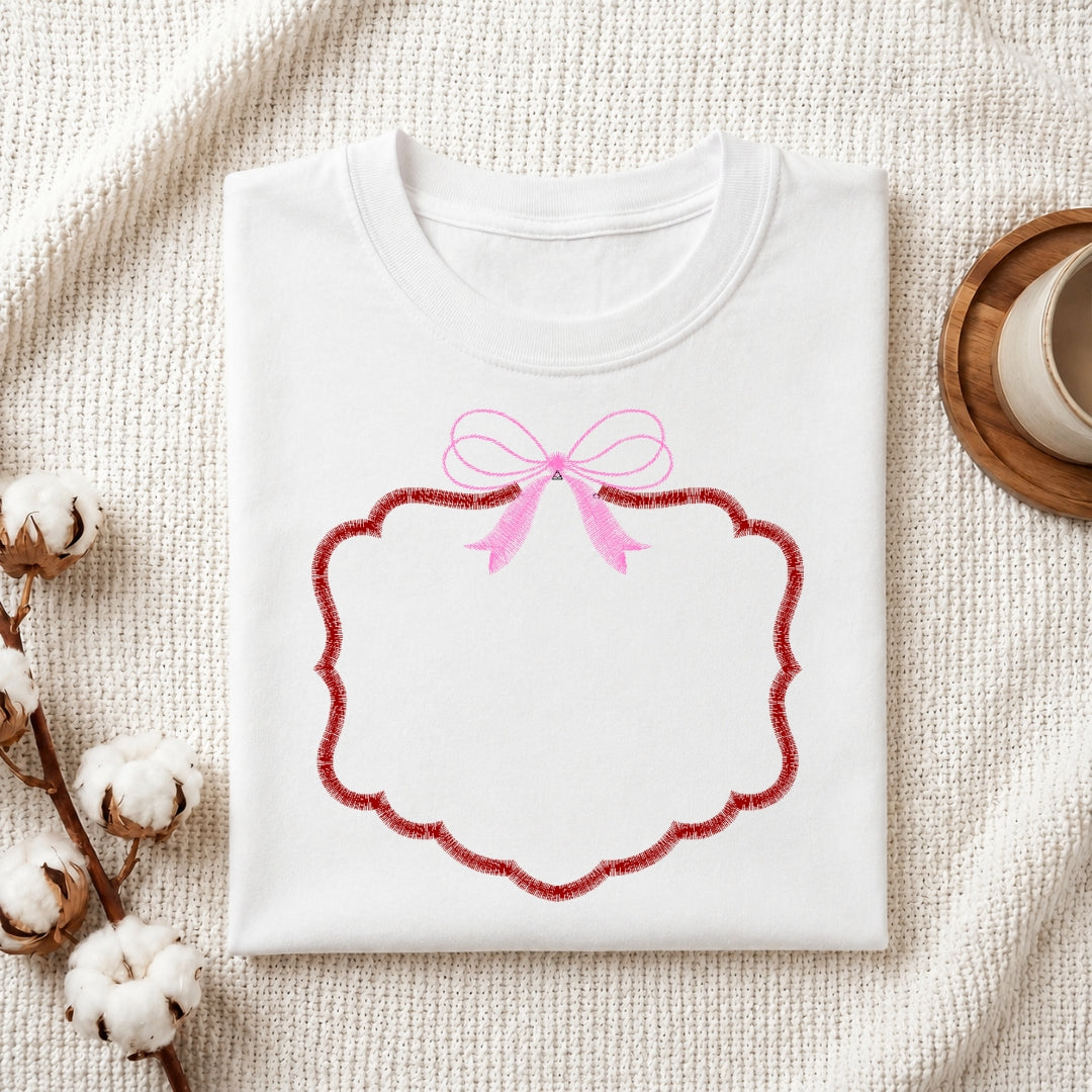 A folded white crewneck sweatshirt with a pink bow and a red scalloped decorative frame graphic on the front, styled on a textured cream fabric with cotton bolls and a wooden cup.