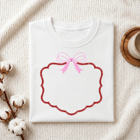A folded white crewneck sweatshirt with a pink bow and a red scalloped decorative frame graphic on the front, styled on a textured cream fabric with cotton bolls and a wooden cup.