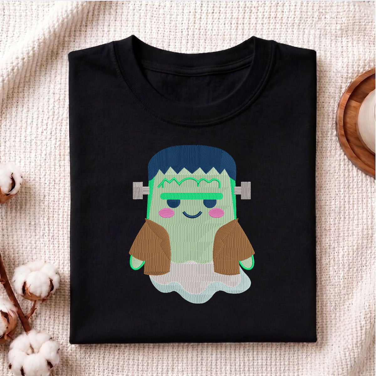 A folded black t-shirt featuring a cute, cartoon-style graphic of Frankenstein's monster with green skin, blue hair, and brown sleeves, displayed on a textured beige fabric background with decorative cotton bolls.