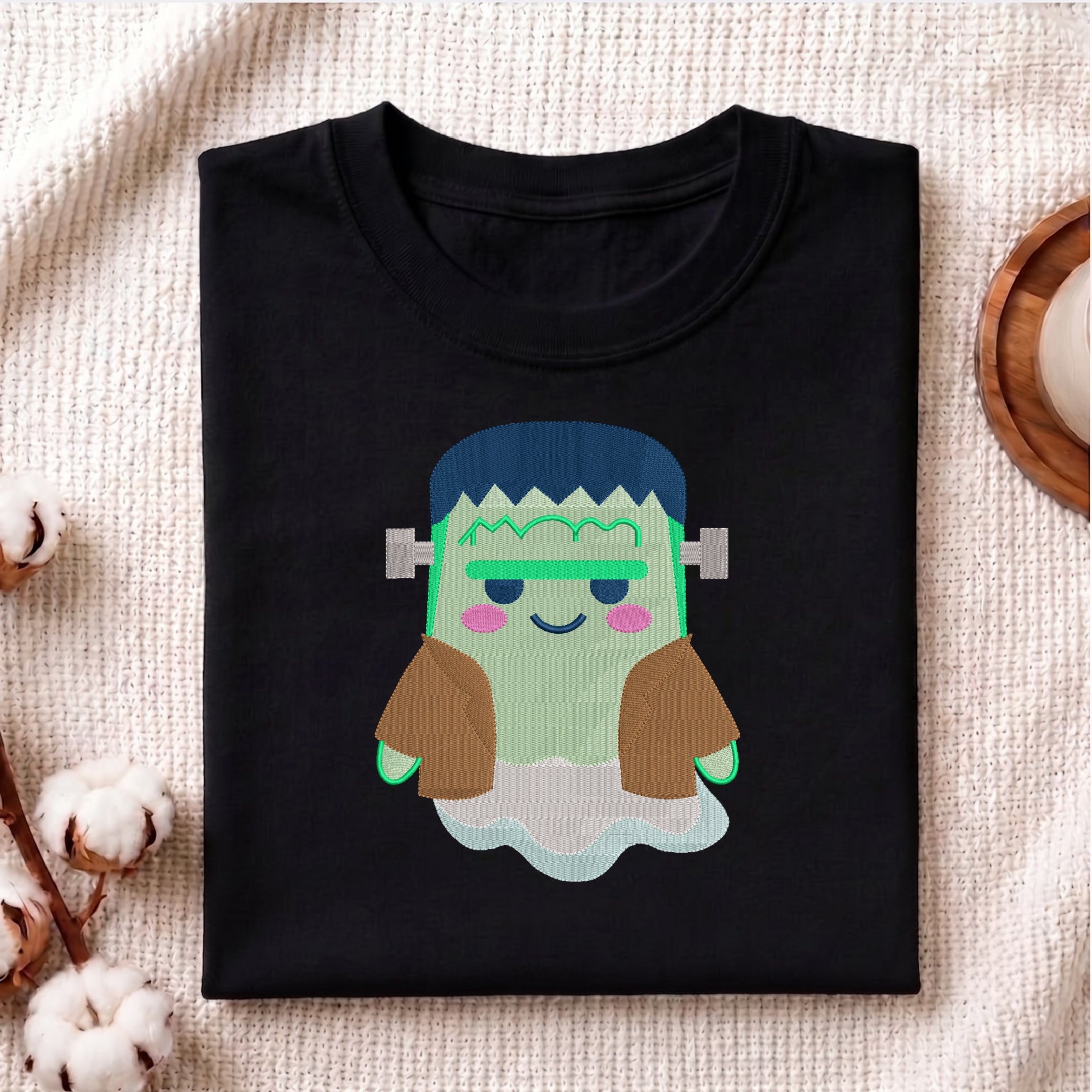 A folded black t-shirt featuring a cute, cartoon-style graphic of Frankenstein's monster with green skin, blue hair, and brown sleeves, displayed on a textured beige fabric background with decorative cotton bolls.
