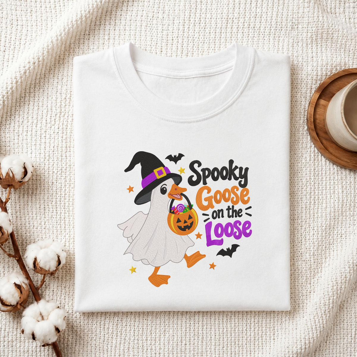 Folded white cotton t-shirt mockup featuring a "Spooky Goose on the Loose" machine embroidery design, styled with cotton stems and a wooden cup