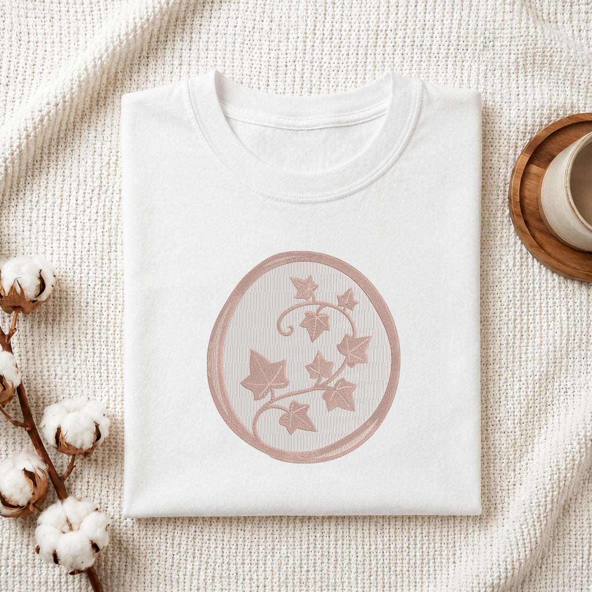 A folded white crewneck t-shirt with a dusty rose circular graphic of ivy leaves, styled on a textured cream blanket with cotton bolls and a wooden cup in the background.
