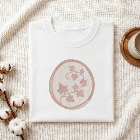 A folded white crewneck t-shirt with a dusty rose circular graphic of ivy leaves, styled on a textured cream blanket with cotton bolls and a wooden cup in the background.