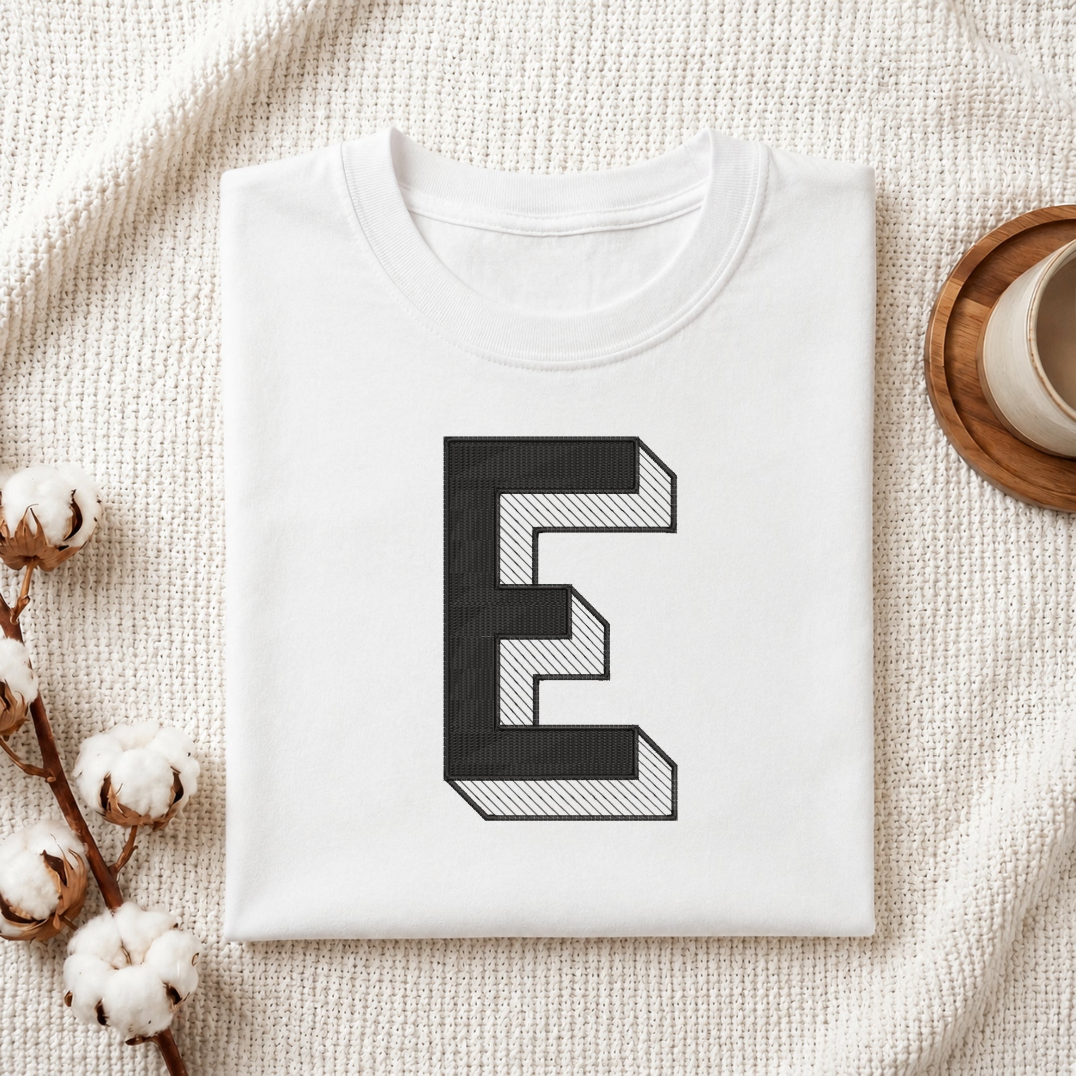 A folded white T-shirt with a large black-and-white geometric letter 'E' printed on the front, styled on a textured cream fabric with cotton stems and a small cup in the background.