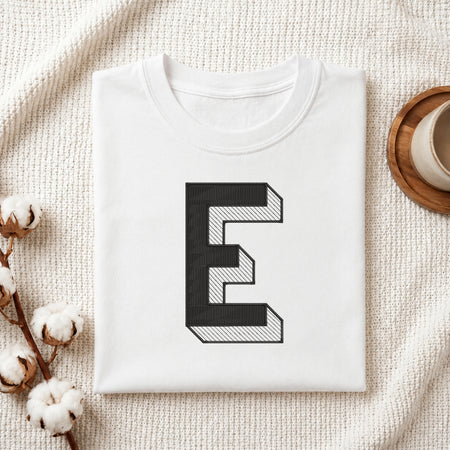 A folded white T-shirt with a large black-and-white geometric letter 'E' printed on the front, styled on a textured cream fabric with cotton stems and a small cup in the background.