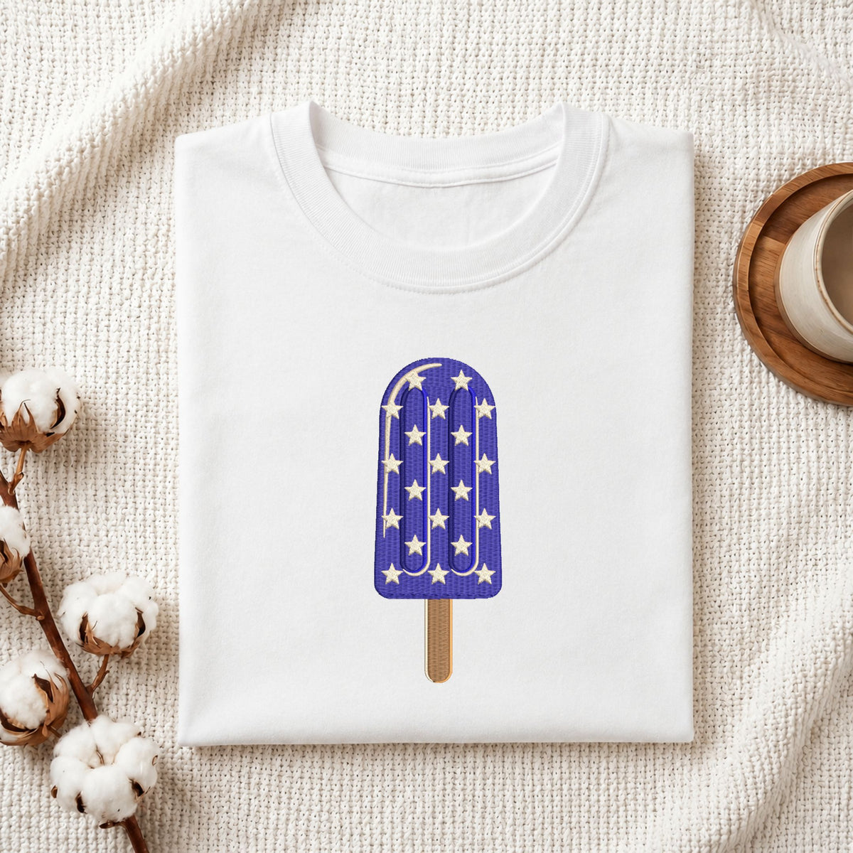 Folded white cotton t-shirt mockup featuring a purple star pattern ice cream popsicle machine embroidery design, styled with cotton stems and a wooden cup.