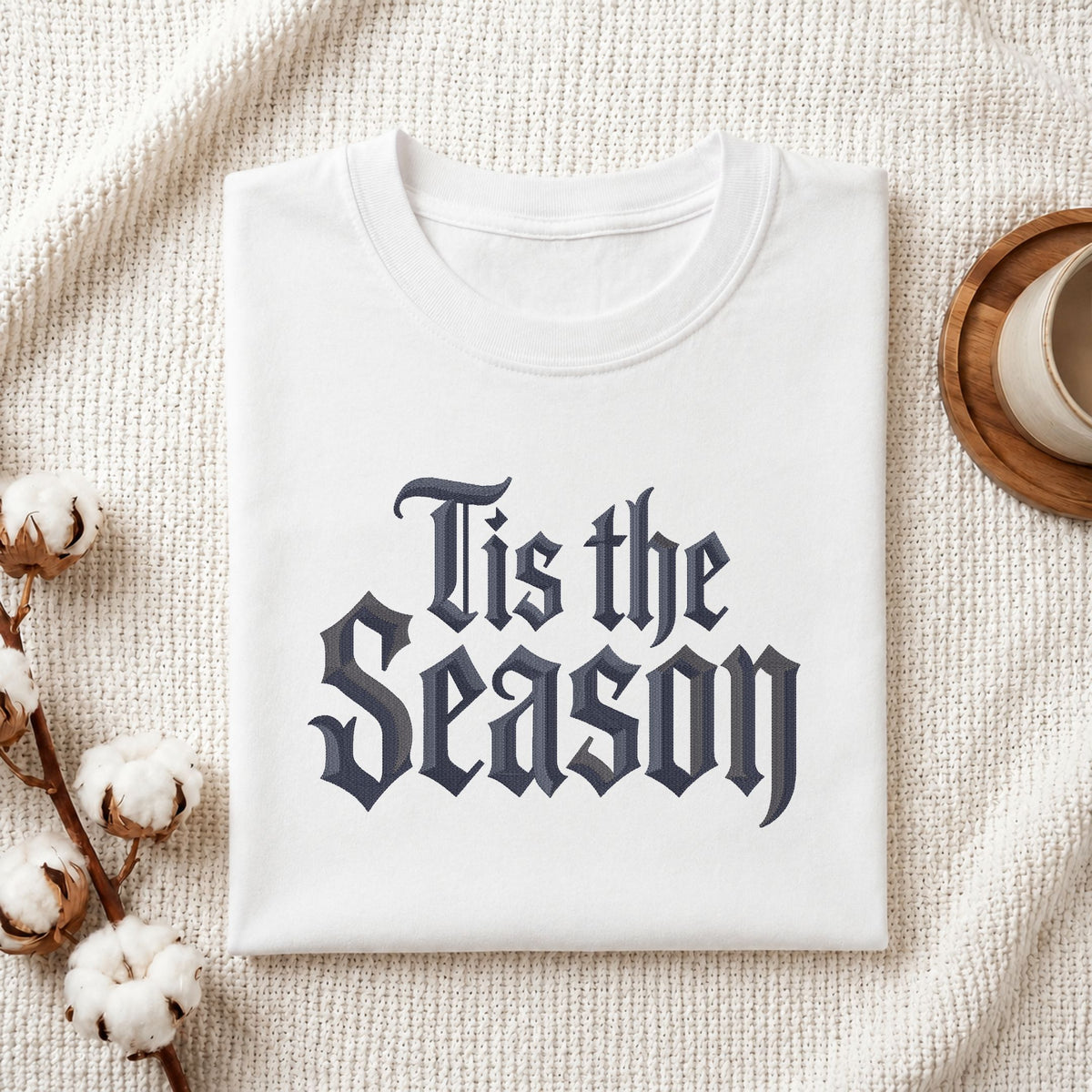 A folded white crewneck t-shirt with the phrase 'Tis the Season' printed in a dark navy gothic font, styled on a textured cream knit blanket with cotton bolls and a wooden cup in the background.