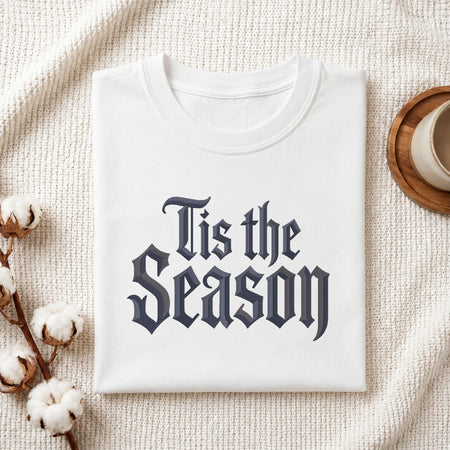 A folded white crewneck t-shirt with the phrase 'Tis the Season' printed in a dark navy gothic font, styled on a textured cream knit blanket with cotton bolls and a wooden cup in the background.