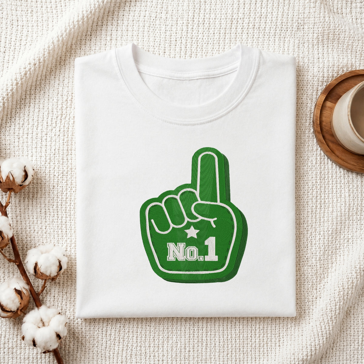 A folded white t-shirt with a green foam finger graphic that says 'No.1' in white, styled on a textured cream fabric with cotton bolls and a wooden cup.