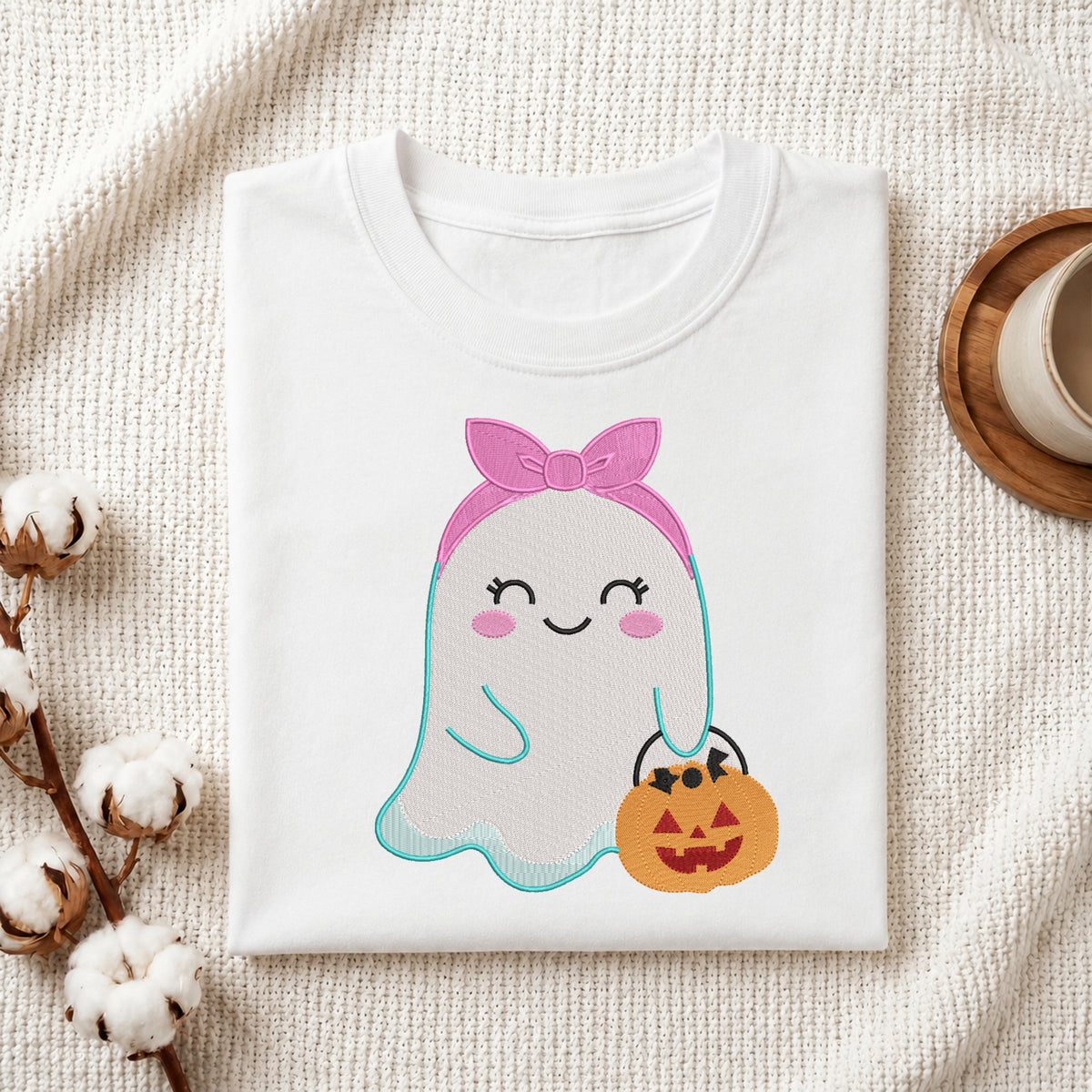 A folded white t-shirt with a cute cartoon ghost graphic holding a jack-o'-lantern bucket, styled on a textured cream fabric with cotton bolls and a coffee cup.