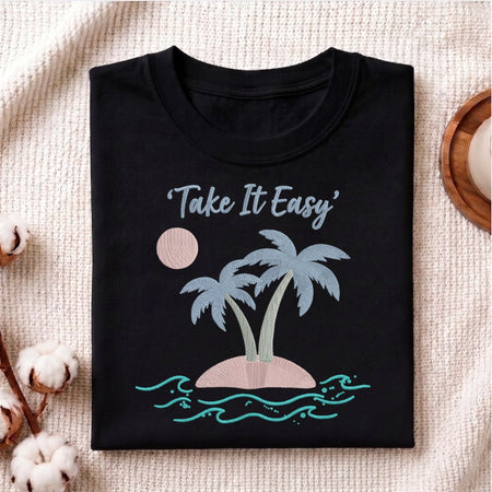 A folded black t-shirt with a 'Take It Easy' tropical island graphic, displayed on a textured beige fabric background with decorative cotton bolls.
