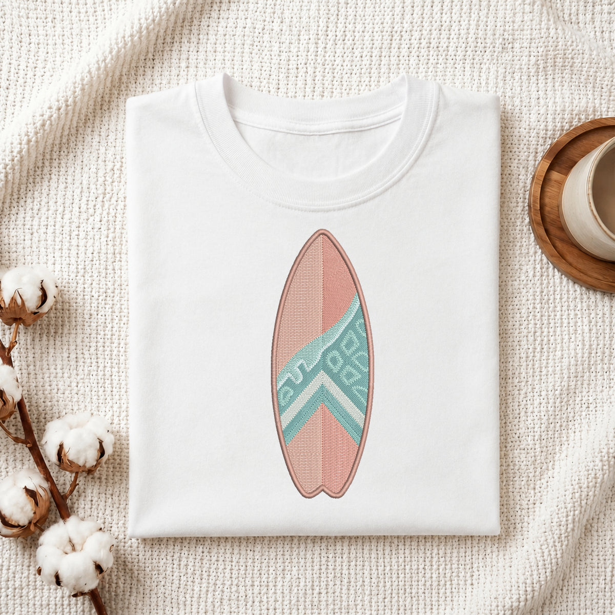 A folded white crewneck t-shirt featuring a printed surfboard design in pink and teal, styled on a textured cream fabric with cotton bolls and a wooden cup.
