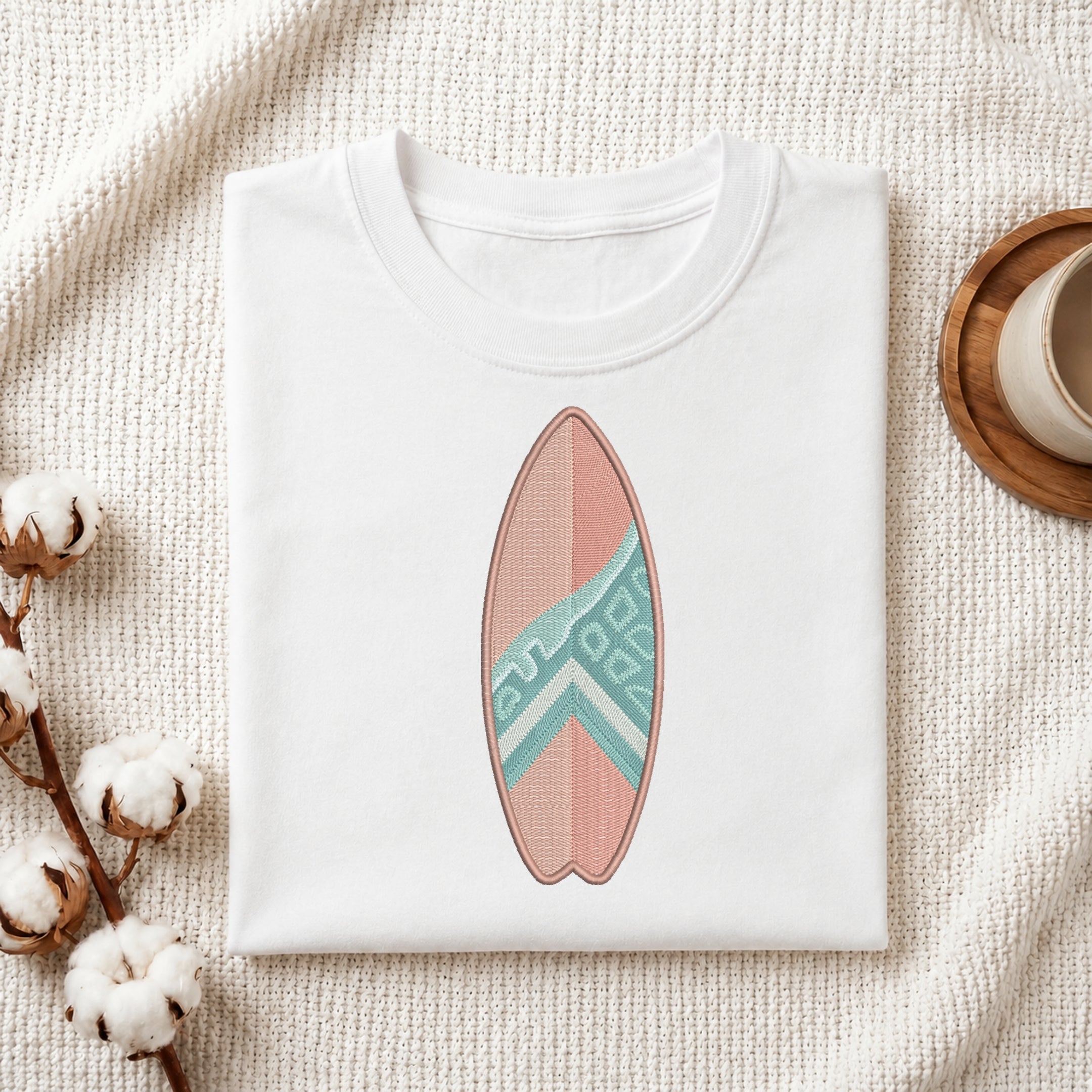 A folded white crewneck t-shirt featuring a printed surfboard design in pink and teal, styled on a textured cream fabric with cotton bolls and a wooden cup.