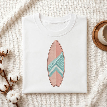A folded white crewneck t-shirt featuring a printed surfboard design in pink and teal, styled on a textured cream fabric with cotton bolls and a wooden cup.