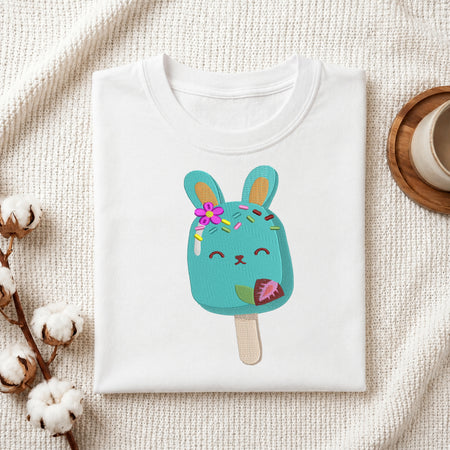 A folded white crewneck t-shirt featuring a cute turquoise bunny popsicle graphic with a pink flower and sprinkles, styled on a textured cream knit blanket with cotton bolls and a wooden cup in the background.