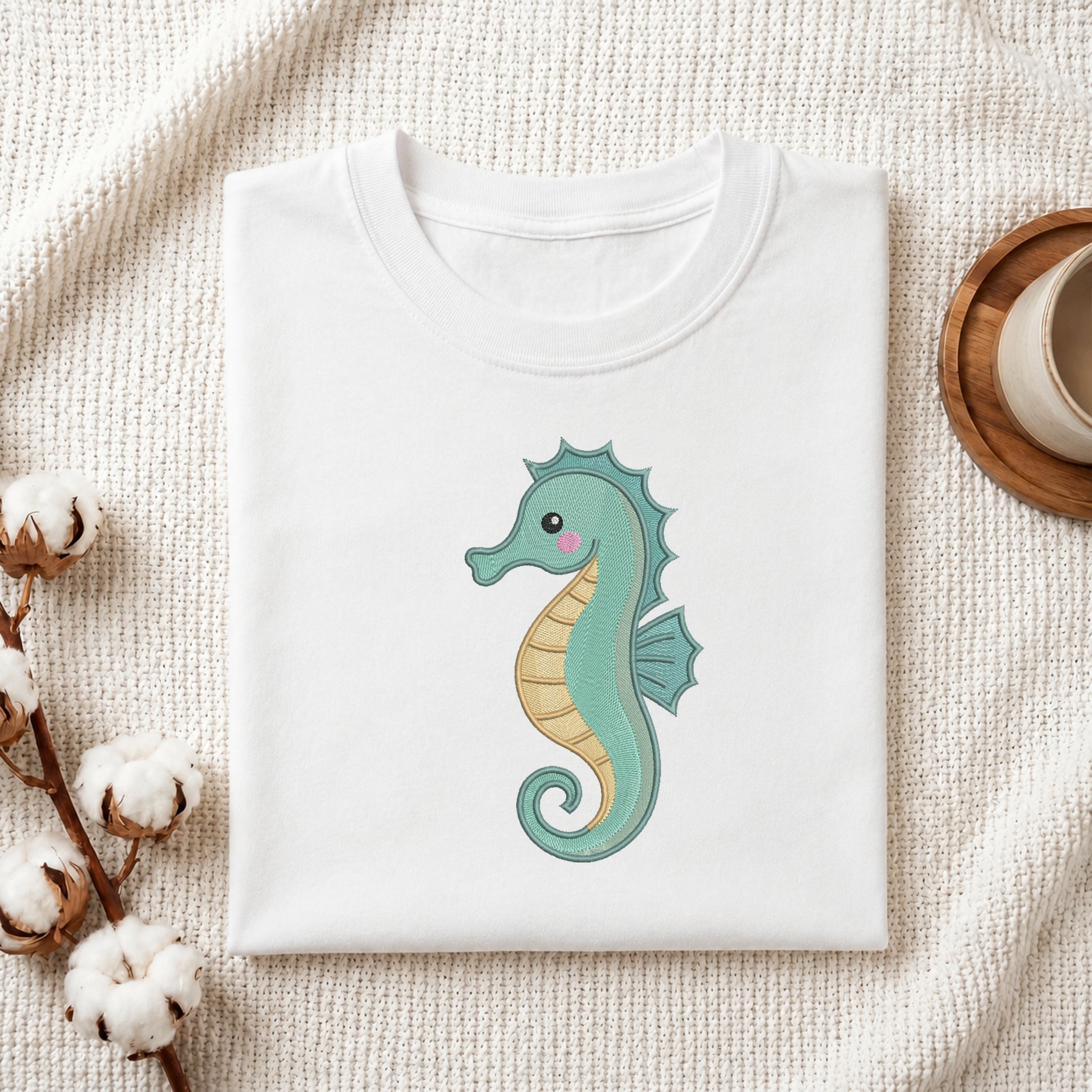 A folded white crewneck t-shirt featuring a cute, embroidered-style seahorse graphic in mint green and pale yellow, displayed on a textured cream fabric background with decorative cotton bolls and a wooden cup.