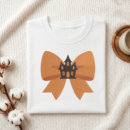 A neatly folded white t-shirt featuring the embroidered orange bow and haunted house design, displayed on a textured white knit background with cotton stems and a wooden cup.