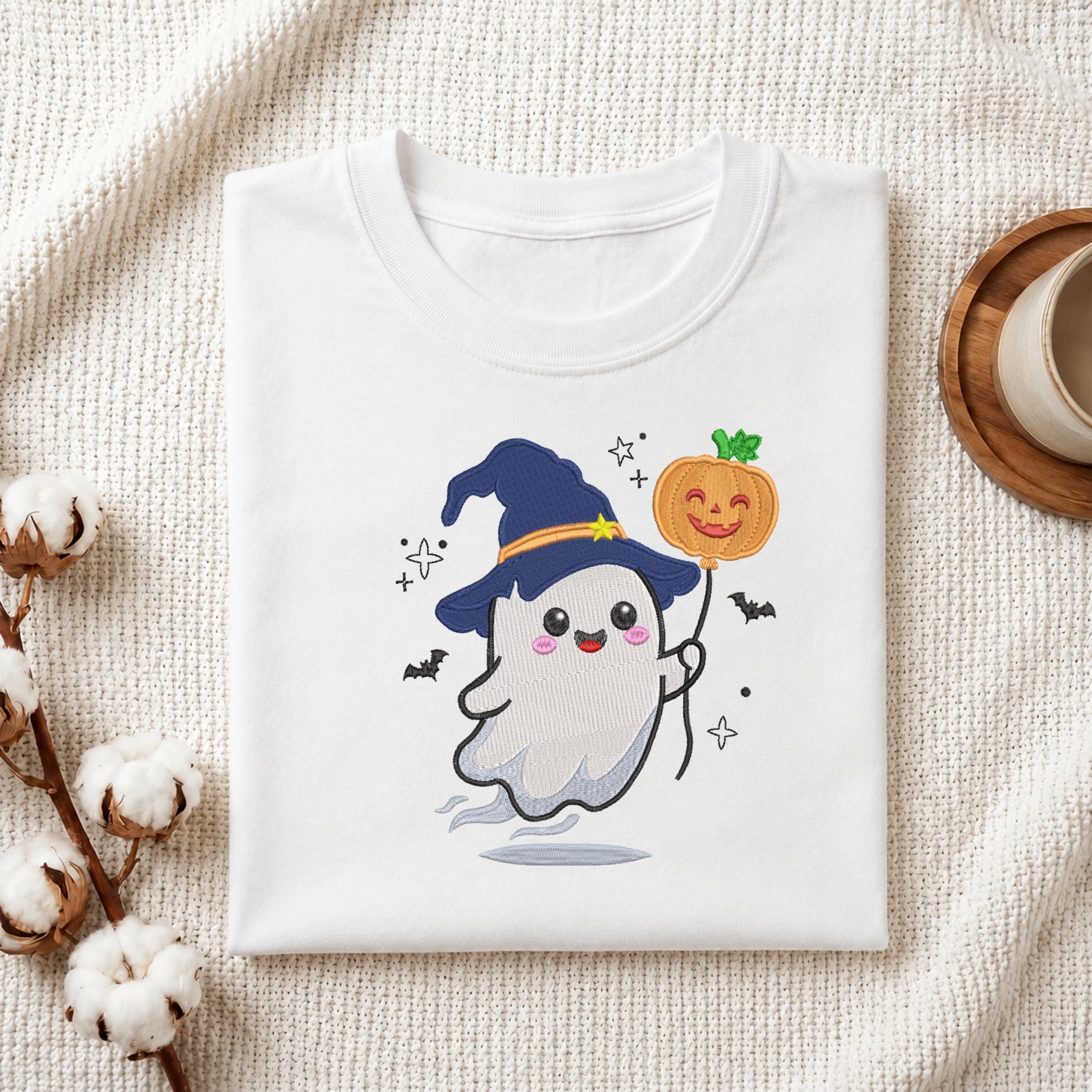 A folded white T-shirt with a cute cartoon ghost wearing a blue witch hat and holding a smiling orange pumpkin, styled on a textured cream fabric with cotton stems and a cup in the background.