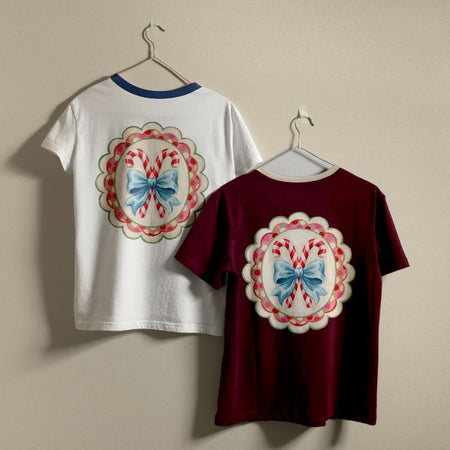 Two short-sleeve crewneck T-shirts hanging on hangers against a plain beige wall. The left shirt is white with a blue collar trim and a large circular candy cane graphic on the back.