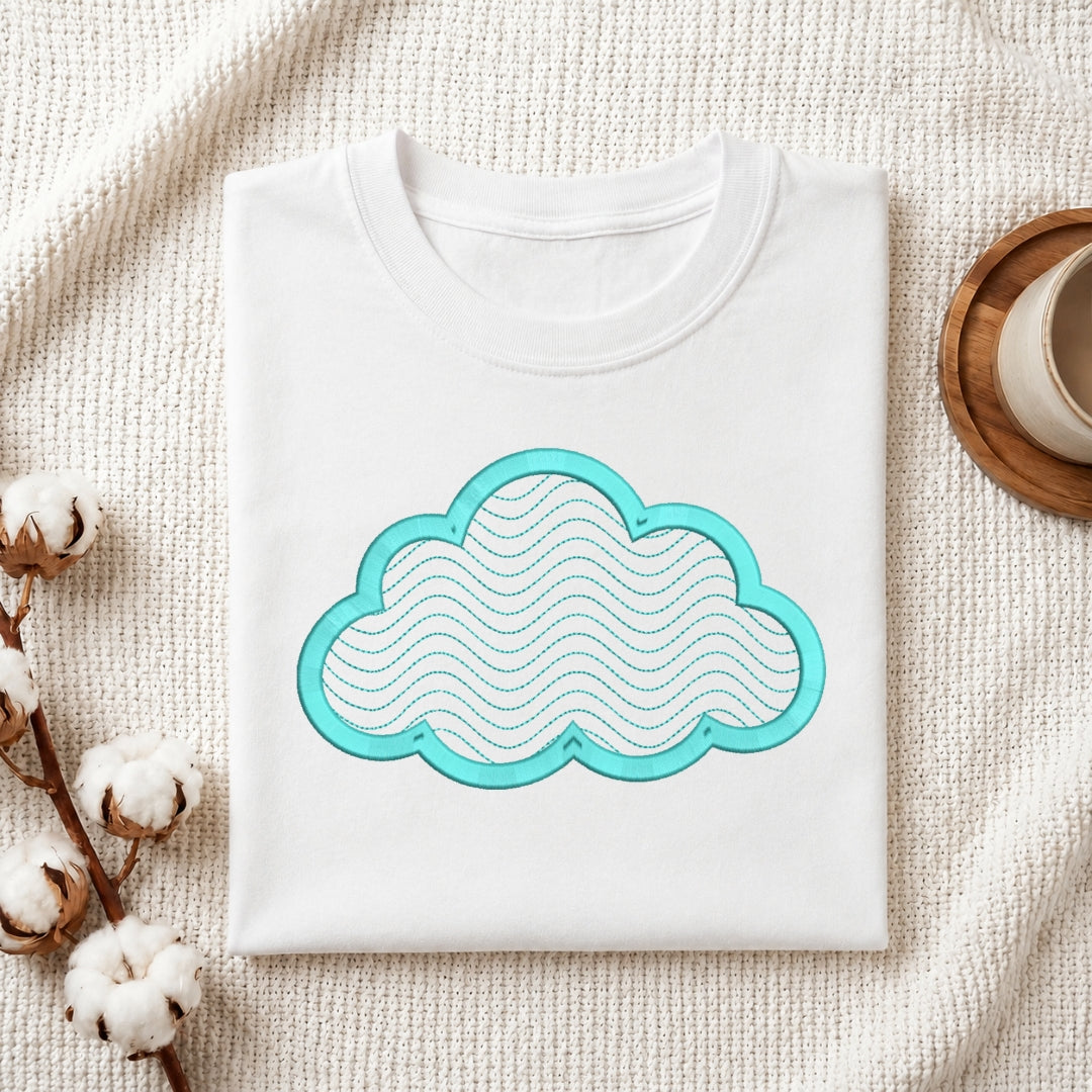 A folded white crewneck t-shirt with a turquoise cloud graphic on the front, styled on a textured cream knit blanket with cotton bolls and a wooden cup in the background.
