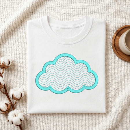 A folded white crewneck t-shirt with a turquoise cloud graphic on the front, styled on a textured cream knit blanket with cotton bolls and a wooden cup in the background.