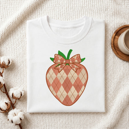 A folded white crewneck t-shirt featuring a graphic of a strawberry with a pink and beige argyle pattern and green leaves, styled on a textured cream fabric with cotton bolls and a wooden cup.