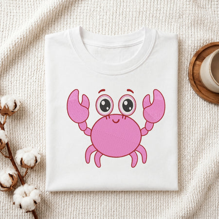 A folded white crewneck t-shirt featuring a cute pink cartoon crab graphic on the front, styled on a textured cream knit blanket with cotton bolls and a wooden cup in the background.