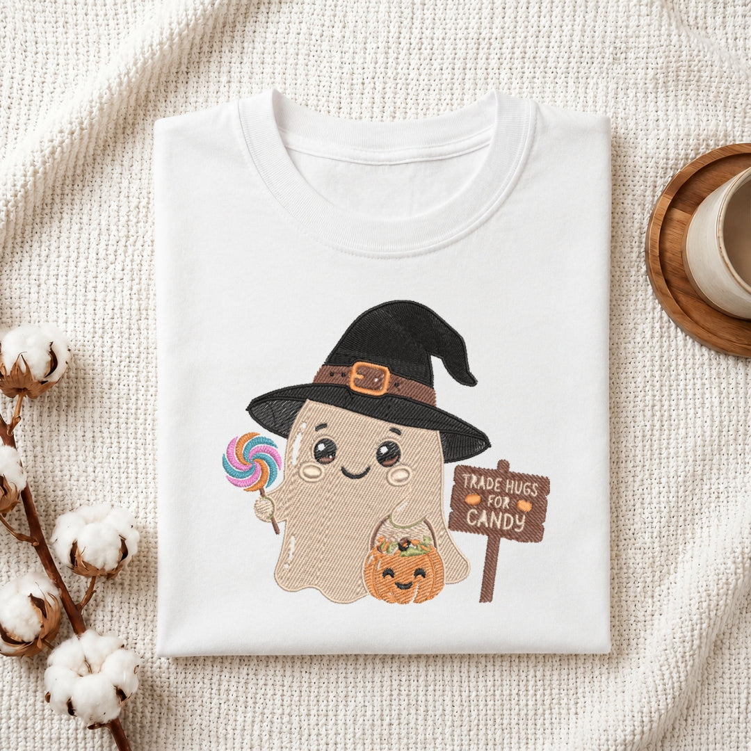 A folded white t-shirt with a cute cartoon ghost wearing a black witch hat, holding a lollipop and a pumpkin, next to a sign that says 'TRADE HUGS FOR CANDY'. The t-shirt is styled on a textured cream fabric with cotton bolls and a cup on a wooden tray.