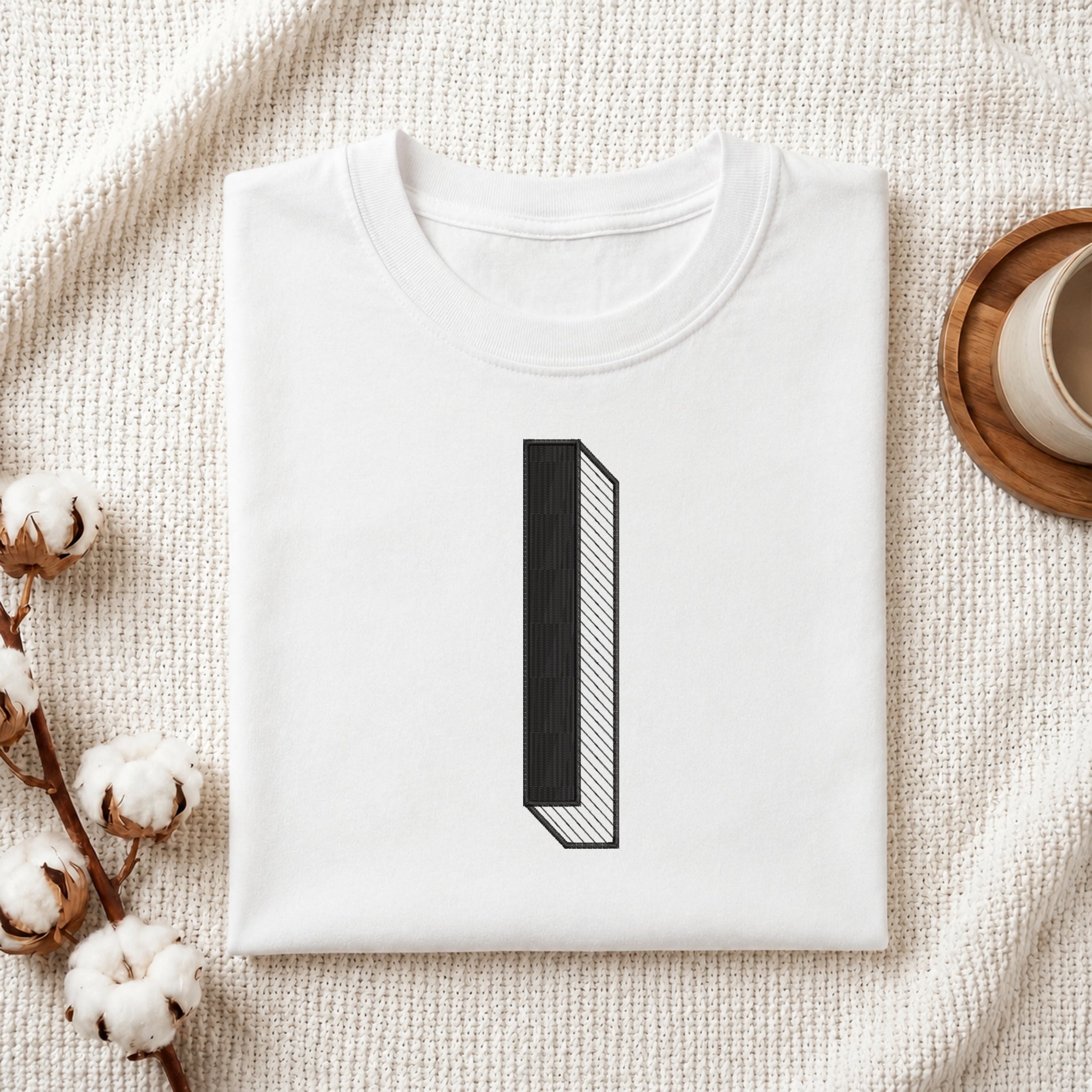 A folded white t-shirt with a minimalist black graphic of the number one, styled on a textured cream blanket with cotton bolls and a wooden cup in the background.