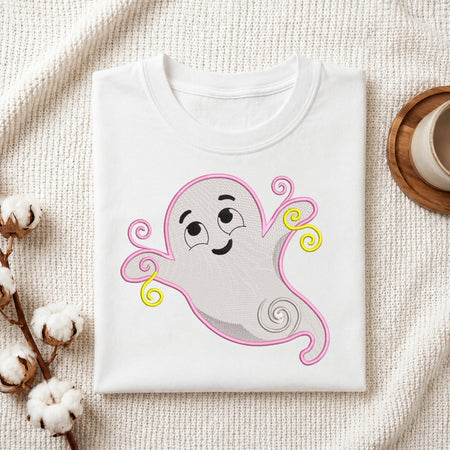 A folded white T-shirt or sweatshirt laid flat on a textured cream fabric background, featuring a cartoon ghost graphic with pink outline, gray body, black facial features, and yellow accents. Cotton stems and part of a cup on a coaster are visible as styled props.